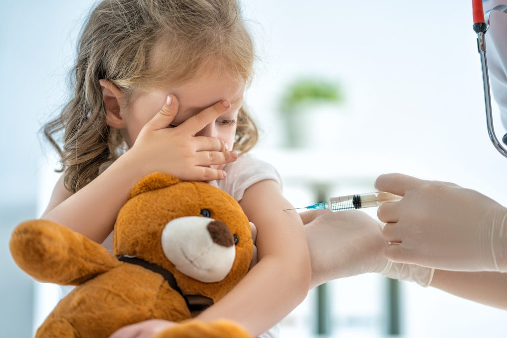 A child holding a teddy bear and covering their eyes while receiving a vaccination.